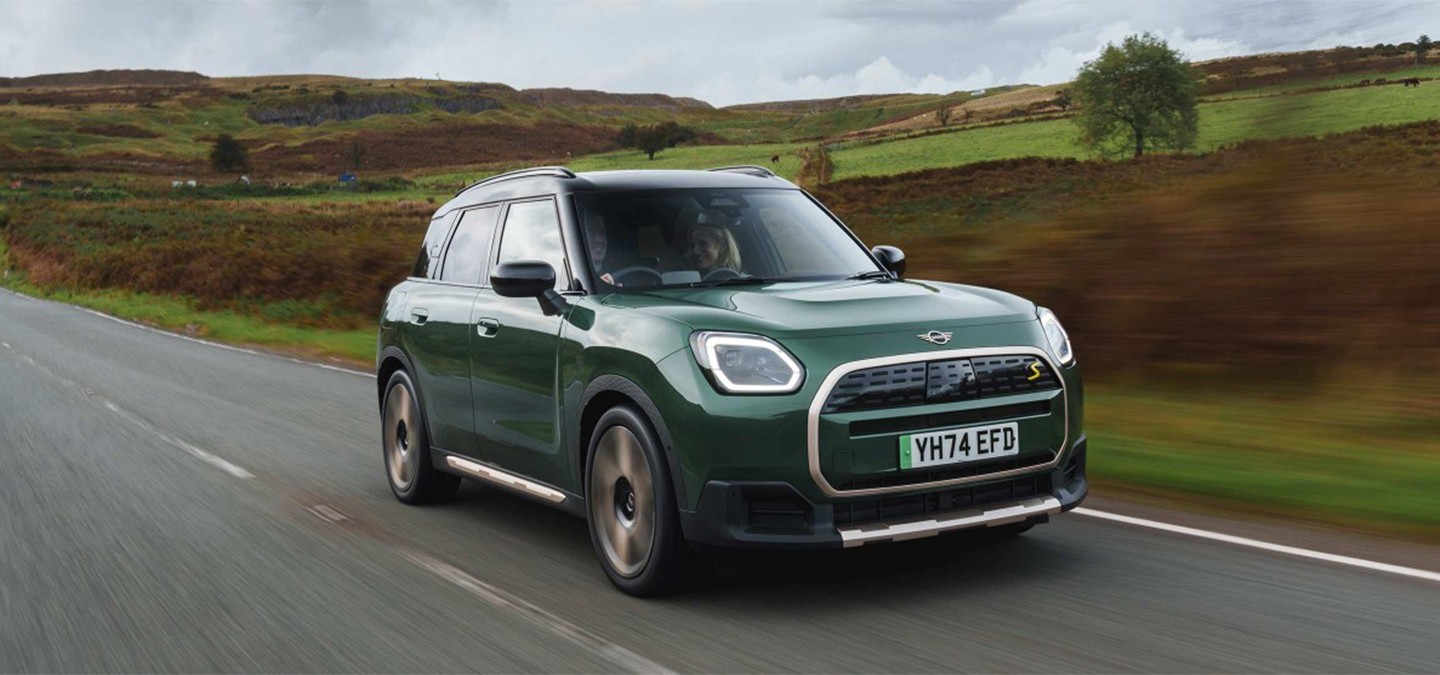 Side view of a driving all-electric MINI Countryman in Smokey Green with Vibrant Silver Roof off-road.